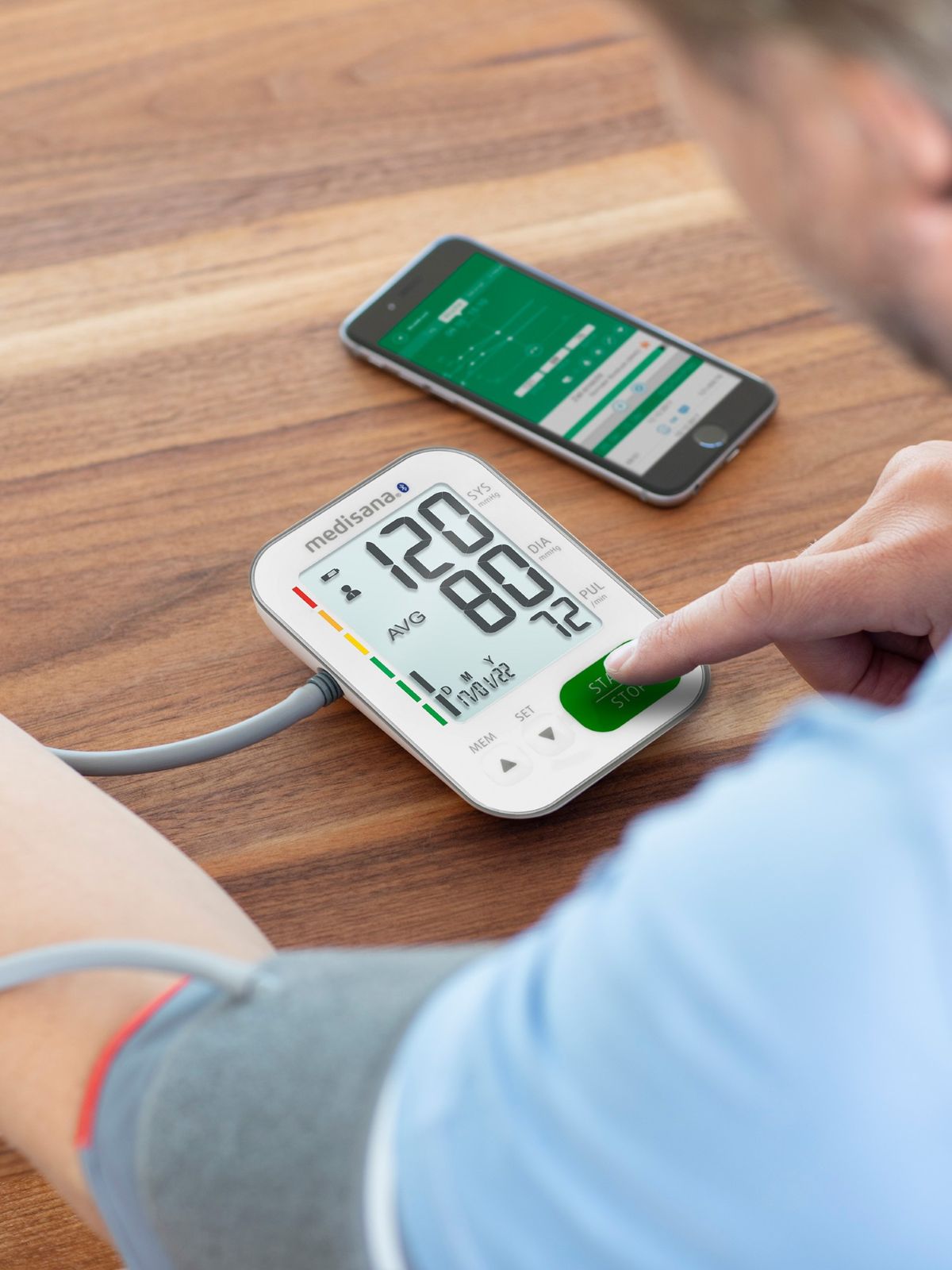 Eine Person misst ihren Blutdruck mit einem digitalen Oberarm-Blutdruckmessgerät, während neben dem Gerät ein Smartphone mit passender Gesundheits-App liegt.